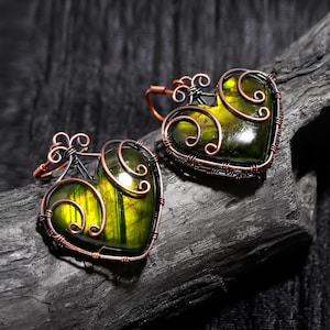 May include: Two heart-shaped earrings with a green and yellow stone center, framed by copper wire and decorative swirls. The earrings are set against a dark, textured background, highlighting the intricate wirework and the vibrant colors of the stones.