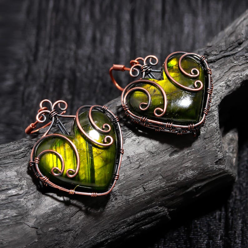 May include: Two heart-shaped earrings with a green and yellow stone center, framed by copper wire and decorative swirls. The earrings are set against a dark, textured background, highlighting the intricate wirework and the vibrant colors of the stones.