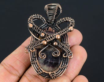May include: A handcrafted pendant featuring an oval-shaped, dark-colored stone encased in intricate copper wire. The wirework includes decorative loops and swirls, creating a unique, artistic design. The pendant is held in a hand against a dark background.