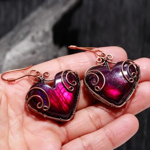 Puede incluir: Un par de pendientes en forma de corazón con un vibrante centro de piedra preciosa púrpura y rosa. Las piedras preciosas están encapsuladas en alambre de cobre con remolinos decorativos. Los pendientes tienen ganchos para las orejas de cobre.