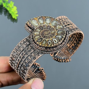 May include: Copper-coloured cuff bracelet with a large, fossilised ammonite centre. The ammonite shows a spiral pattern in shades of brown and tan. The bracelet has a woven wire design.