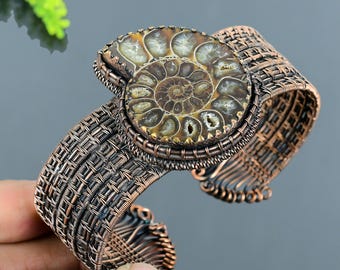 May include: Copper-coloured cuff bracelet with a large, fossilised ammonite centre. The ammonite shows a spiral pattern in shades of brown and tan. The bracelet has a woven wire design.