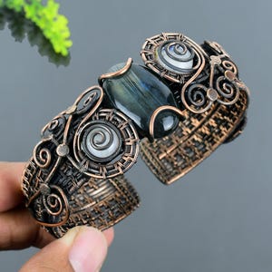 May include: A handcrafted copper cuff bracelet featuring a central oval-shaped, dark blue-green gemstone. The bracelet has intricate wirework with spiral and circular designs, and a woven texture. The overall design is ornate and artistic.