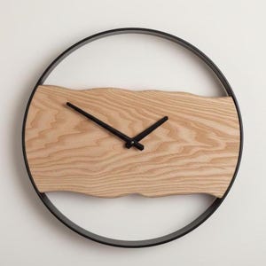 Puede incluir: Un reloj de pared moderno con una esfera de madera clara y agujas negras. La esfera, de forma rectangular con borde superior ondulado, está enmarcada en un círculo negro. Las agujas son negras.