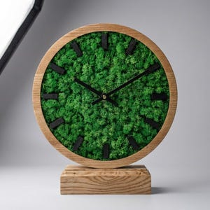 May include: A round wooden clock with a natural moss face and black hands and markers. The clock is set on a wooden base. The clock's face is filled with vibrant green moss, creating a unique and natural aesthetic. The clock measures approximately 8 inches in diameter.