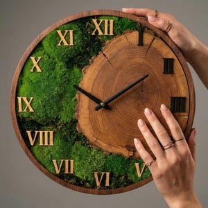 Op de afbeelding: Een ronde houten wandklok met een natuurlijk houtnerfcentrum en een omringende rand. De wijzerplaat heeft groen mos en gouden Romeinse cijfers. Zwarte wijzers geven de tijd aan. De klok heeft een diameter van ongeveer 30 cm.