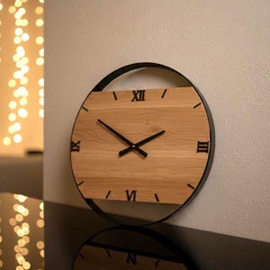 Puede incluir: Un reloj de pared redondo con una esfera de madera marrón claro y números romanos negros. El reloj tiene manecillas negras y un marco de metal negro. El reloj está montado en una pared blanca.