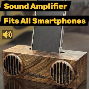 Puede incluir: Un soporte para teléfono y amplificador de sonido rectangular de madera de nogal natural con dos rejillas de altavoz circulares y una ranura central para un teléfono inteligente. El texto dice: Phone Stand, Sound Amplifier, Fits All Smartphones, Radiophone, Natural Walnut Wood.