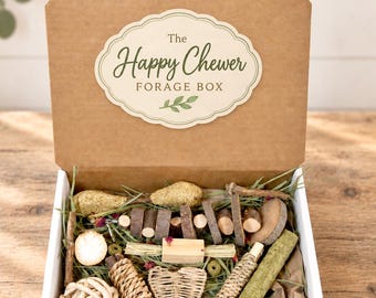 May include: A brown cardboard box with an open lid featuring a label that reads The Happy Chewer FORAGE BOX. Inside are various natural items including a woven heart, wooden sticks, a ball of twine, and green forage material for small animals.