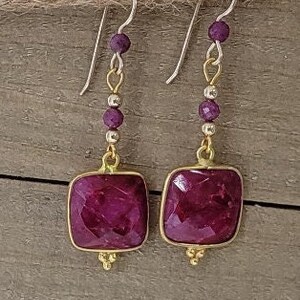 May include: A pair of dangle earrings with square-shaped purple gemstones and gold accents. The earrings have a silver chain with small purple beads.