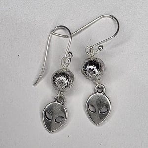 May include: A pair of silver-toned earrings. Each earring features a textured ball and a small alien head charm with oval eyes. The earrings have a hook closure. The background is a plain white surface.