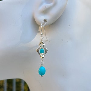Op de afbeelding: Zilveren oorhangers met turquoise stenen. De oorhangers hebben een geometrisch ontwerp met een kleinere turquoise steen in het midden en een grotere turquoise steen die eronder hangt.
