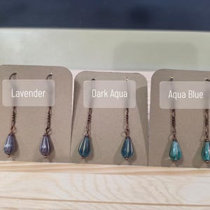 May include: Earrings in three colors: lavender, dark aqua, and aqua blue. Each earring features a teardrop-shaped bead suspended from a copper-colored chain. The earrings are displayed on individual cardboard backings.