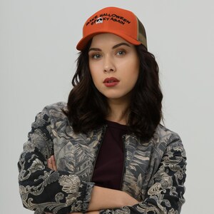 May include: An orange and khaki trucker hat with the text "MAKE HALLOWEEN SPOOKY AGAIN" in black. The hat features a skull graphic and a curved brim. The hat is worn by a person.