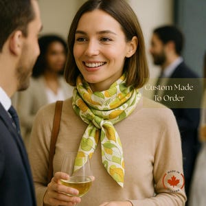 May include: A woman wearing a beige sweater and a silk scarf with a leaf pattern in green, yellow, and white. The scarf is knotted at the neck. The text "Custom Made To Order" is visible. A glass of wine is held in the hand.