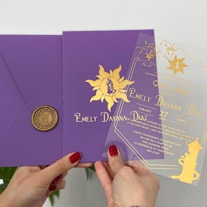 Puede incluir: Invitación de boda morada con detalles dorados. La invitación presenta un emblema de sol y los nombres "Emely Dayana Diaz". Se ve una superposición transparente con texto dorado y diseños florales.