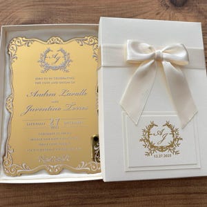 Op de afbeelding: Een trouwuitnodiging in een cr&egrave;mekleurige doos met een satijnen lint en strik. De gouden uitnodigingskaart heeft sierlijke details en de namen "Andrea Laralle" en "Juventino Torres", met de datum "12.27.2025".