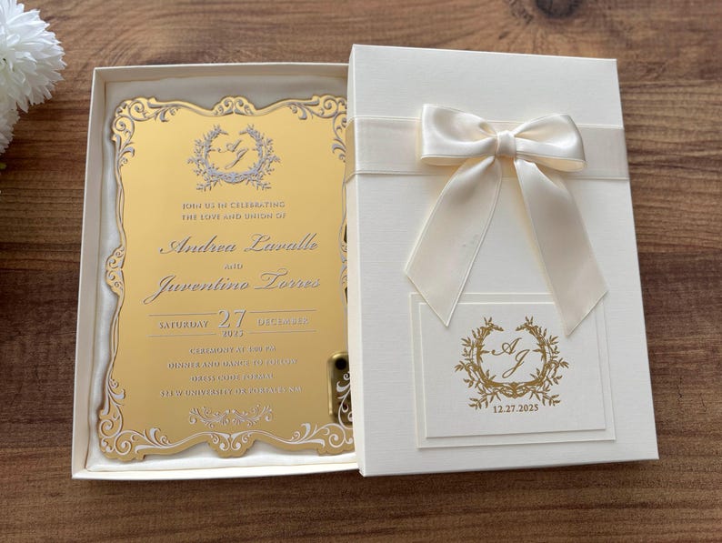 Op de afbeelding: Een trouwuitnodiging in een cr&egrave;mekleurige doos met een satijnen lint en strik. De gouden uitnodigingskaart heeft sierlijke details en de namen "Andrea Laralle" en "Juventino Torres", met de datum "12.27.2025".