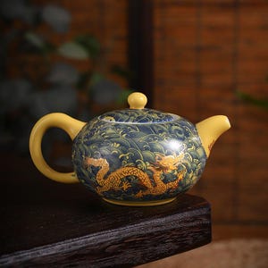 May include: A ceramic teapot with a yellow handle, spout, and lid. The body of the teapot is blue with a wave pattern and a gold dragon design. The teapot is sitting on a dark wooden surface.