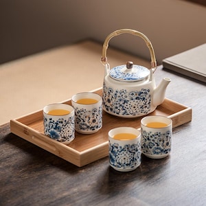 Może przedstawiać: Biały ceramiczny zestaw do herbaty z niebieskimi kwiatowymi wzorami, w tym czajniczek z bambusowym uchwytem i cztery pasujące filiżanki wypełnione herbatą. Zestaw stoi na prostokątnej bambusowej tacy.