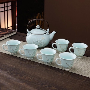 Puede incluir: Un juego de té de cerámica azul claro con una tetera y seis tazas a juego. La tetera tiene un asa de metal y detalles florales. Las tazas y la tetera tienen un delicado diseño floral. El juego se exhibe sobre una superficie de madera.