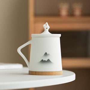 Puede incluir: Taza de cerámica blanca con tapa y asa, con un diseño de montaña en tonos grises. La taza se apoya sobre una base de bambú. La tapa tiene un asa decorativa. La taza está sobre una superficie blanca.