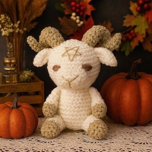 Pode incluir: Boneca de ovelha de crochê feita à mão, creme, com chifres, cascos e detalhes faciais bege. Uma estrela dourada é costurada na testa. Duas abóboras laranja estão em ambos os lados do carneiro. O fundo inclui folhagem de outono.