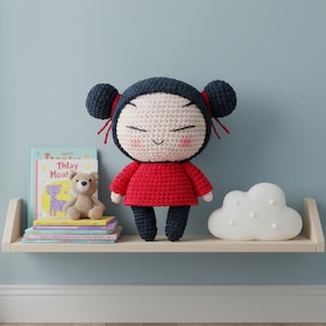 May include: A handmade crochet doll with a red dress, black pants, and dark blue hair buns tied with red ribbons. The doll has a smiling face with closed eyes. A small teddy bear and books are on a shelf below.