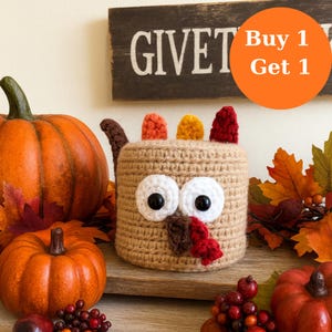 May include: A crocheted turkey-shaped container with large white eyes and a red wattle. The container is surrounded by pumpkins, fall leaves, and berries. A wooden sign that says "GIVE" is in the background. An orange circle with the text "Buy 1 Get 1" is also in the background.