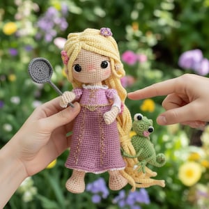 Könnte beinhalten: Handgefertigte Häkelpuppe einer Prinzessin mit langem blondem Haar, die ein rosa Kleid trägt und einen grauen Gegenstand hält. Ein kleiner grüner Frosch befindet sich neben der Puppe. Die Puppe befindet sich vor einem verschwommenen Hintergrund aus Blumen.
