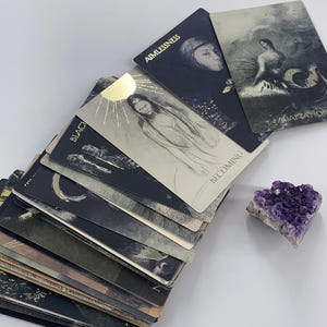May include: A deck of oracle cards with illustrations in shades of grey and black, featuring words like "Aimlessness" and "Becoming." A purple amethyst crystal sits nearby. The cards have a mystical theme.