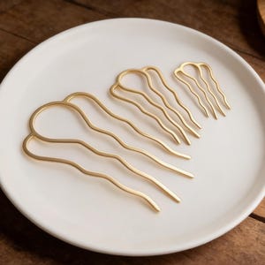 May include: Four gold-colored hair pins in varying sizes arranged on a white ceramic plate. The pins have a wavy design. The plate sits on a dark wooden surface, with a small wooden dish and twine in the background.