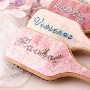 Peut inclure: Quatre brosses à cheveux personnalisées avec des manches en bois et des dessus roses, blancs et crème. Chaque brosse présente un nom épelé en strass étincelants. Les noms sont Rachel, Vivienne, Shirley et Natalie. Le fond comprend des fleurs violettes.