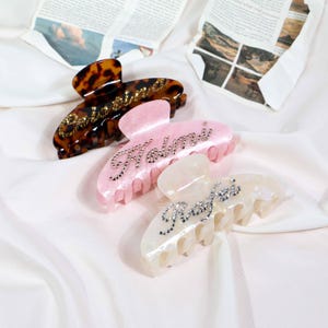 May include: Three hair claw clips in the image. One is tortoiseshell with gold-colored text, another is pink with silver text, and the last is white with silver text. The text on each clip spells out a name.