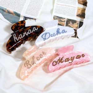 May include: Four hair claw clips in various colors: tortoiseshell, white, peach, and pink. Each clip features a name spelled out in rhinestones. The names are: "hanaa", "Duba", "Belooa", and "Mayar".