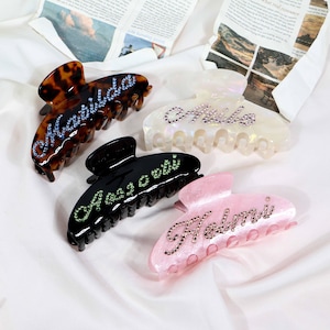 May include: Four hair claw clips in various colors: tortoiseshell, iridescent white, black, and pink. Each clip features a name spelled out in sparkling rhinestones. The names are Matilda, Mila, Alessia, and Halmi.