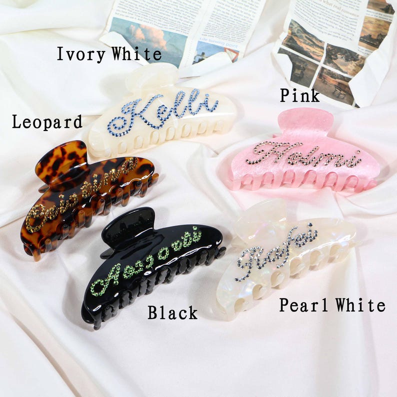 May include: Five hair claw clips in various colors: ivory white, leopard print, black, pink, and pearl white. Each clip features a name spelled out in rhinestones. The clips are arranged on a white, silky surface.