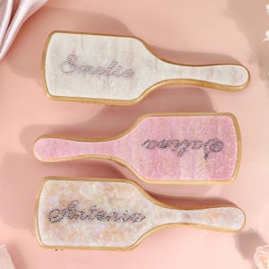 May include: Three personalized hairbrushes with wooden handles and pearlescent tops. The brushes are in shades of white, pink, and cream, each with a name spelled out in rhinestones. The names are "Emelie", "Dakota", and "Antonia".