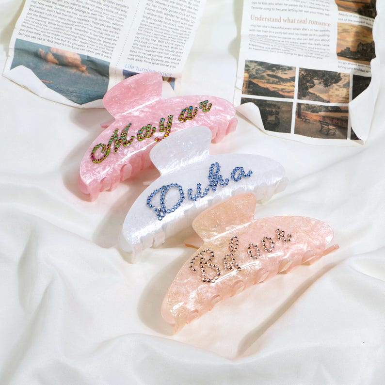 May include: Three acrylic hair claw clips in pink, white, and peach. Each clip has a name spelled out in rhinestones: "Mayar", "Duka", and "Belove". The clips are on a white, textured surface.