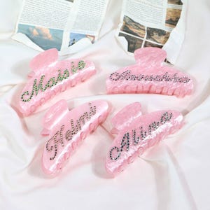 May include: Four pink hair claw clips with names spelled out in rhinestones. The names are "Maisie", "Anouschka", "Helmi", and "Alivia". The clips are on a white satin surface.