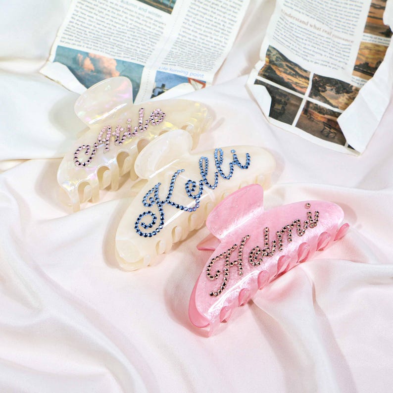 May include: Four personalized hair claw clips. Three are pearlescent white with names spelled out in rhinestones: "Amile", "Kelli", and "Helmi". The fourth clip is pink with the name "Helmi" in rhinestones. The clips are on a white satin surface.