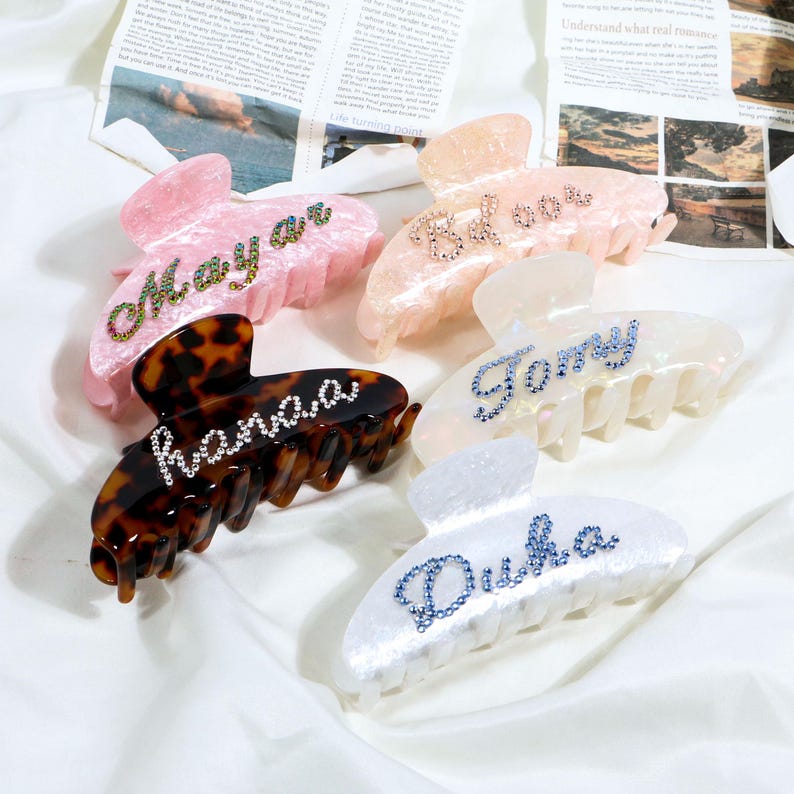 May include: Five claw hair clips in various colors: pink, peach, tortoiseshell, white, and iridescent white. Each clip features a name spelled out in rhinestones. The names are: Mayar, Belove, hana.a, Tony, and Duka.