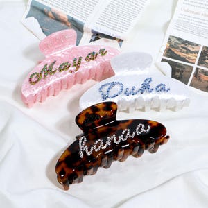 May include: Three hair claw clips in pink, white, and tortoiseshell patterns. Each clip features a name spelled out in rhinestones: "Maya", "Duha", and "Hanaaa". The clips are on a white surface with newspaper in the background.