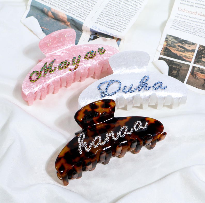 May include: Three hair claw clips in pink, white, and tortoiseshell patterns. Each clip features a name spelled out in rhinestones: "Maya", "Duha", and "Hanaaa". The clips are on a white surface with newspaper in the background.