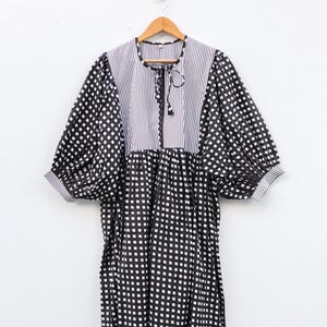 Puede incluir: Vestido a cuadros blancos y negros con corpiño y mangas a rayas, colgado de una percha de madera. El vestido tiene un diseño suelto y fluido con mangas cortas y abullonadas y un lazo en el escote. La tela parece ligera.