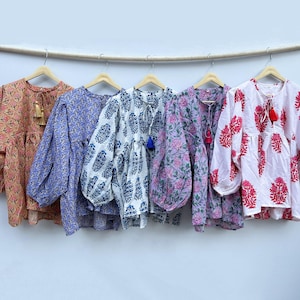 Könnte beinhalten: Fünf langärmlige Blusen mit floralen Mustern hängen an Holzbügeln. Die Blusen sind in den Farbtönen Pfirsich, Lavendel, Weiß, Rosa und Weiß mit roten floralen Designs gehalten. Jede Bluse hat eine Quaste am Ausschnitt.