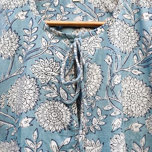 May include: A light blue top with a white floral pattern and a tie-up neckline. The fabric features a repeating design of large flowers and leaves. The top is hanging on a wooden hanger.