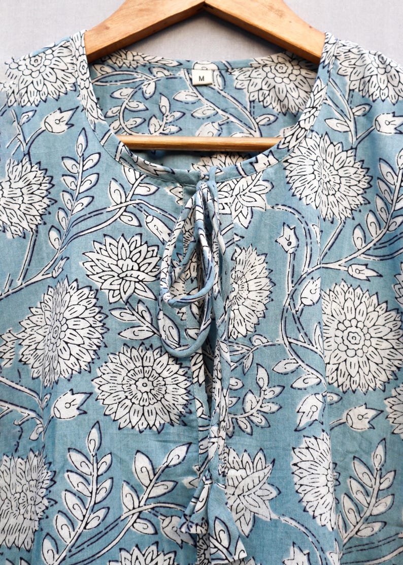 May include: A light blue top with a white floral pattern and a tie-up neckline. The fabric features a repeating design of large flowers and leaves. The top is hanging on a wooden hanger.