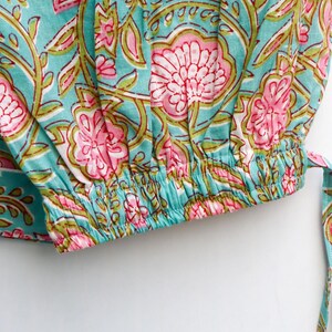 Op de afbeelding: Close-up van een turquoise stof met een bloemenpatroon in roze en groen. Het ontwerp heeft grote, gestileerde bloemen en bladeren. De stof heeft een elastische zoom en een bijpassende stropdas.