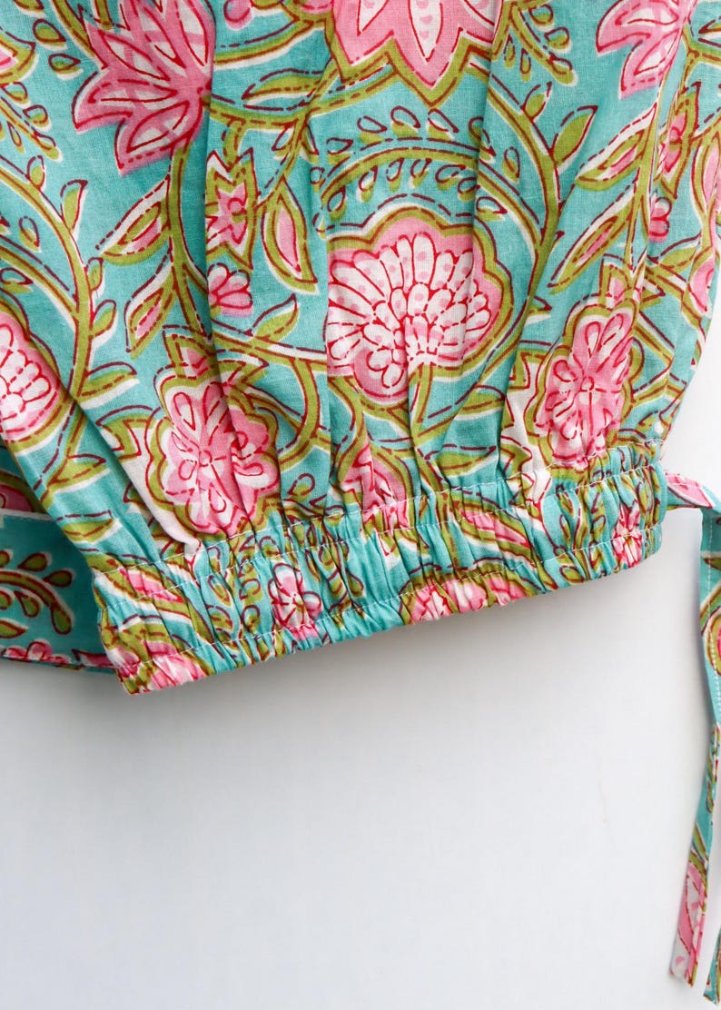 Op de afbeelding: Close-up van een turquoise stof met een bloemenpatroon in roze en groen. Het ontwerp heeft grote, gestileerde bloemen en bladeren. De stof heeft een elastische zoom en een bijpassende stropdas.
